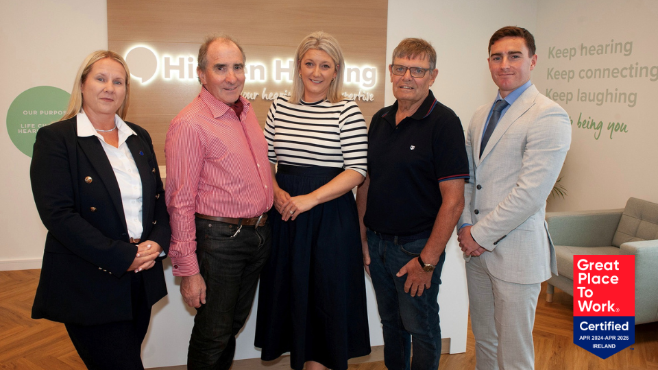 [GEN] Five individuals stand in a professional hearing clinic, in front of a 'Hidden Hearing' sign. Wall text includes: 'Keep hearing, Keep connecting, Keep laughing, Keep being you' and 'Our Purpose: Life-changing hearing care.' A certification badge reads: 'Great Place to Work Certified APR 2024–APR 2025 IRELAND.'