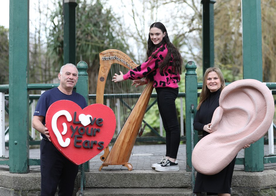 A man holds a red heart-shaped sign reading 'Love Your Ears,' a woman plays a wooden harp, and another holds a large ear sculpture in a park gazebo setting.