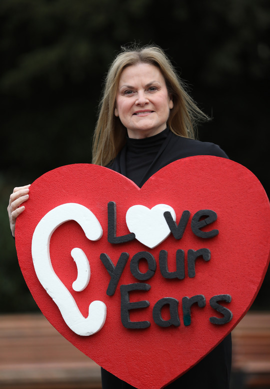 A person holds a large red heart-shaped sign with the text 'Love Your Ears' featuring an ear graphic. The background includes greenery and a blurred park bench, suggesting an outdoor setting.
