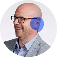 Hidden Hearing audiologist Tom Owens with a big smile while a blue speech bubble is overlayed on his ear