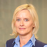 A woman with blonde hair wearing a light blue collared shirt and dark vest is pictured against a blurred neutral background. Her face is obscured for privacy. No text is visible.