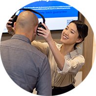 An audiologist places headphones on a man's head during a hearing test in a hearing clinic. A screen in the background displays text, though it's not fully legible.