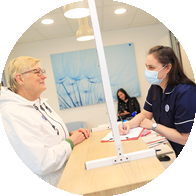 [GEN] A woman in a white hoodie speaks with an audiologist in a navy uniform across a reception desk with a protective screen. The hearing clinic features a modern design with a dandelion-themed blue artwork visible in the background. No specific hearing aid brands are identifiable.