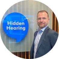 [GEN] A man in a suit stands beside a blue sign featuring the text 'Hidden Hearing' in a professional hearing clinic setting with modern wooden paneling and frosted glass decor elements.