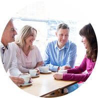 [GEN] Four individuals sit around a wooden table, holding white coffee cups, engaged in a conversation within a bright, casual café setting featuring large windows in the background.