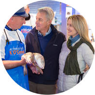 [GEN] A fishmonger holding fresh fish engages with an older man and a woman in a vibrant market setting. The fishmonger wears a blue apron with text saying 'We love what we do.'