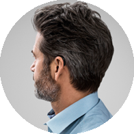 A man with neatly styled dark hair and a trimmed beard is shown in profile, wearing a blue shirt. A discreet, modern hearing aid is visible behind his ear. Context implies professional hearing care.