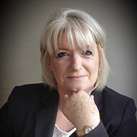 A senior individual with short gray hair rests their hand under their chin, seated in a neutral indoor setting. The face is blurred, obscuring identity. No hearing aids or text are visible.