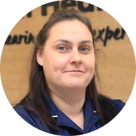 [GEN] A female staff member in a navy uniform, with a blurred face, stands in front of a wooden wall featuring partially visible text: 'Hearing Health... experience.' Likely in a hearing clinic setting.