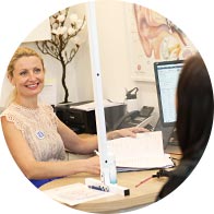 A hearing clinic consultation shows a professional seated behind a desk with papers, assisting a client across from them. The setting includes ear anatomy posters, a computer, and office equipment.