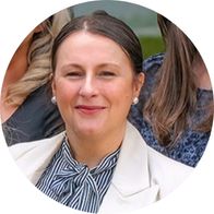 [GEN] A person wearing a white blazer and striped blouse with pearl earrings, seated among others in an outdoor setting with green foliage in the background. The face is blurred. No text present.