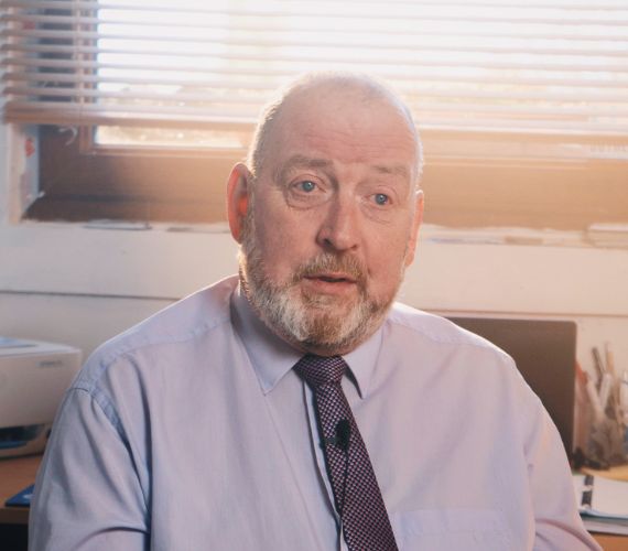 Kevin Quaid in a shirt and tie talking with the sun shining in through the window behind him