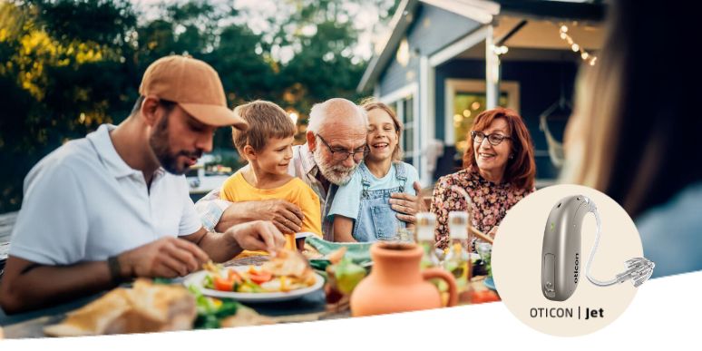 A family sitting around a grandparent at a table eating food outside with the Oticon Jet hearing aid being showcased in a bubble