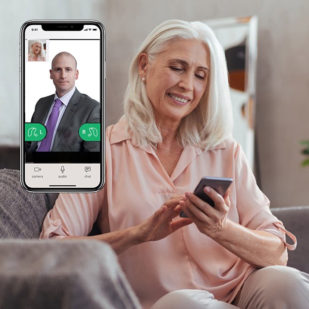 An older woman with white hair holds a smartphone while seated on a sofa in a home setting. The phone displays a video call interface featuring an audiologist wearing a suit and tie. Hearing aid icons for left ('L') and right ('R') ears are visible, along with options labeled 'camera,' 'audio,' and 'chat.'