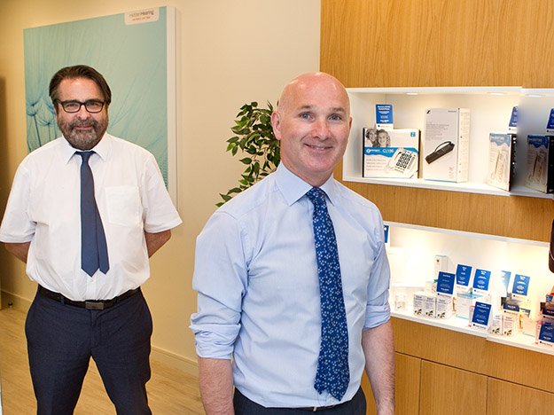 Two men in formal attire stand in a hearing clinic. Display shelves on the right showcase hearing aids, amplifiers, and brochures. A 'Philips' sign is visible in the background.