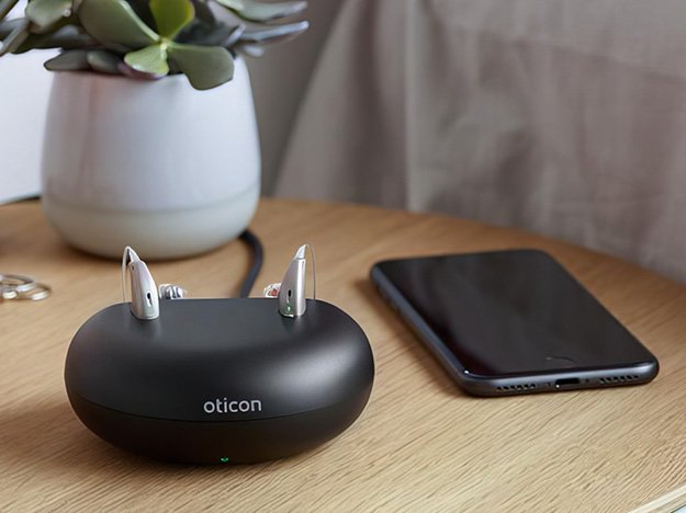 A pair of Oticon hearing aids rest in a sleek black charging case labeled 'Oticon,' sitting on a wooden table beside a smartphone and a potted succulent plant in a cozy indoor setting.