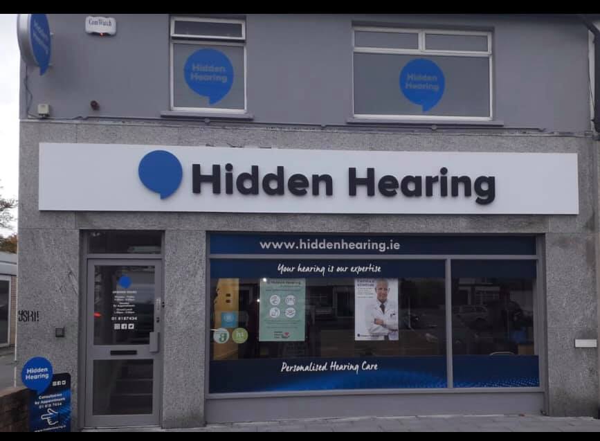 The storefront of Hidden Hearing features bold signage with a blue speech bubble logo and the text 'Hidden Hearing.' The window displays 'www.hiddenhearing.ie,' 'Your hearing is our expertise,' and 'Personalised Hearing Care.' The clinic offers hearing-related services, as indicated by branding for hearing aids. The building exterior is gray with a glass door promoting contact details and services.