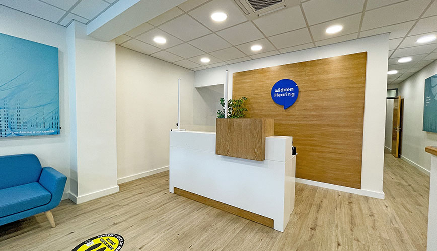 A modern hearing clinic reception desk stands with a wooden accent wall featuring a round blue sign that reads 'Hidden Hearing.' The space includes a blue chair, light wood flooring, and soft lighting for a welcoming environment.