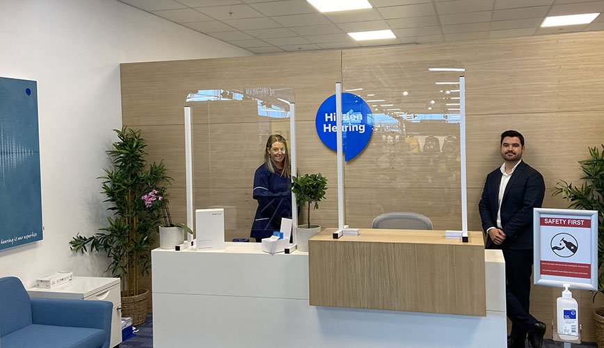 A reception desk at a Hidden Hearing clinic features clear dividers, safety signage, and a branded blue logo. A staff member stands behind the desk, and a man stands nearby. Natural plants, seating, and sanitizer enhance the welcoming and hygienic environment.