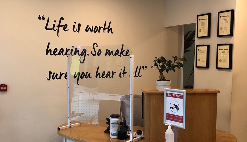 A reception area in a hearing clinic displays the quote, 'Life is worth hearing. So make sure you hear it all,' on a wall. The wooden desk holds a clear protective screen, hand sanitizer, and cleaning wipes. Nearby are framed audiology certificates and a small potted plant, creating a welcoming environment for visitors seeking hearing tests or hearing aids.