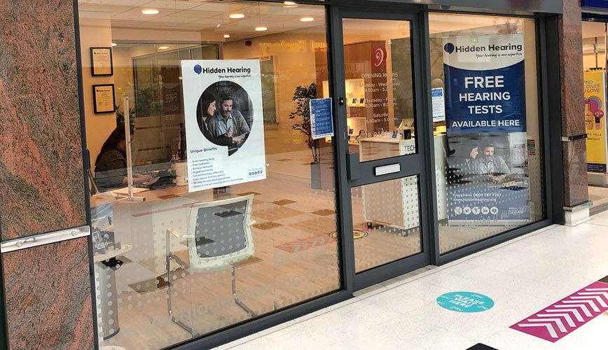 A Hidden Hearing clinic storefront features large glass windows, displaying posters reading 'FREE HEARING TESTS AVAILABLE HERE' and 'Unique Benefits' alongside a logo. The interior shows chairs, counters, and a lit, welcoming environment within a shopping center.
