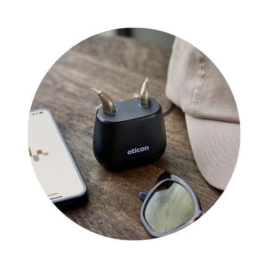 Oticon Intent hearing aids in a charging case placed on a wooden surface alongside a smartphone, sunglasses, and a beige cap