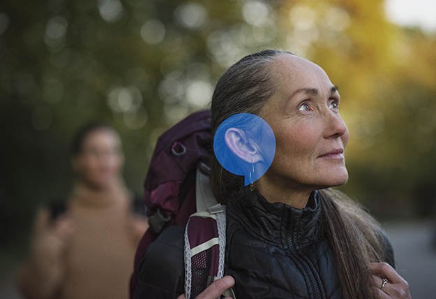 A person wearing outdoor gear and a backpack has a painted blue ear covering their natural ear, standing in a park-like environment with blurred greenery and another individual in the background.