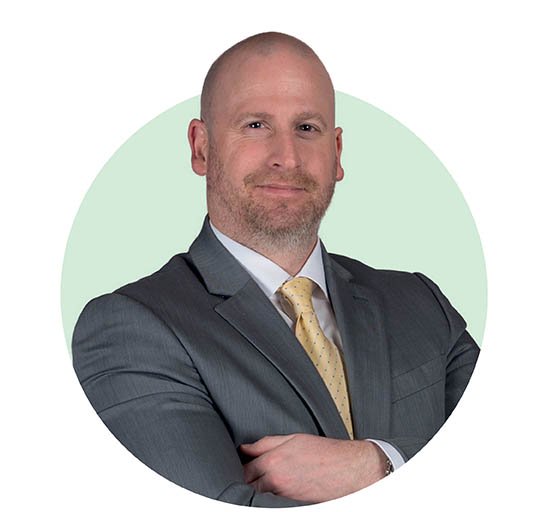 A bald man in a gray suit and yellow tie stands with arms crossed against a pale green circular background. His face is blurred for privacy. No text or hearing-related elements are visible.