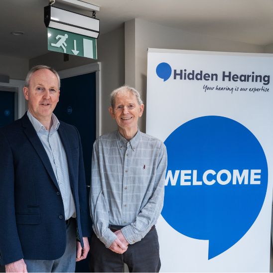 Two men standing while smiling next to a Hidden Hearing sign with a blue speech bubble that says welcome