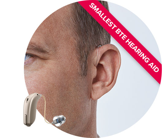 A close-up of a man's ear wearing a discreet behind-the-ear (BTE) hearing aid with a clear tube and earpiece. Text reads: 'SMALLEST BTE HEARING AID.' White background.