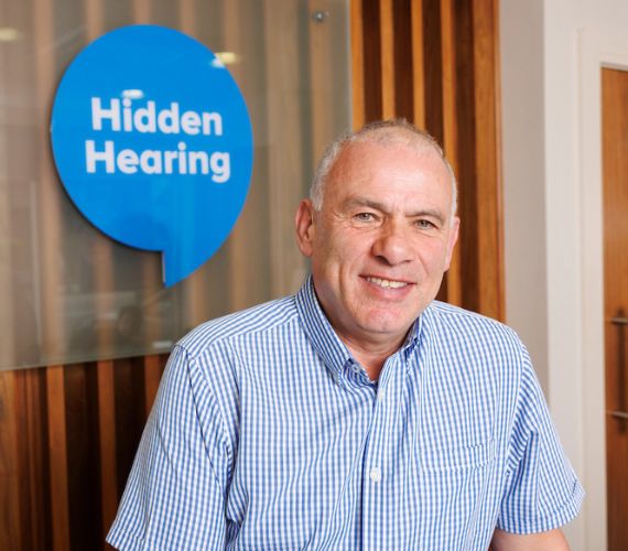 Tony Ward in a blue shirt smiling with a Hidden Hearing speech bubble behind him