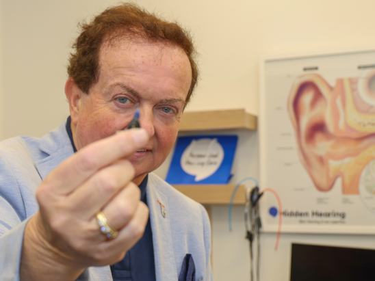 Marty Morrissey holding an Oticon Zeal hearing aid while in a Hidden Hearing clinic