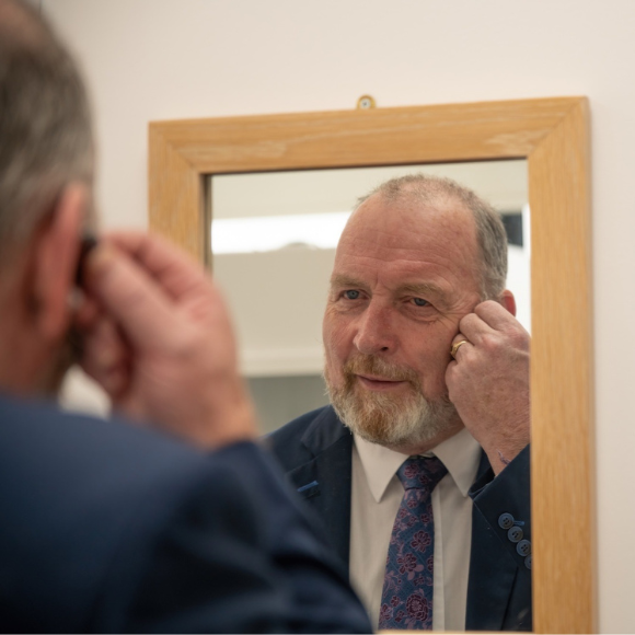 A man in a suit adjusts a hearing aid while looking into a rectangular wooden-framed mirror, positioned in a well-lit indoor environment, likely within a hearing clinic.