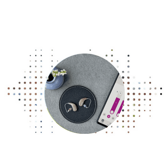 Due apparecchi acustici beige sono posizionati su un elegante supporto rotondo nero, accanto a uno smartphone con un’app di controllo dell’udito e un vaso decorativo in un ambiente minimalista.