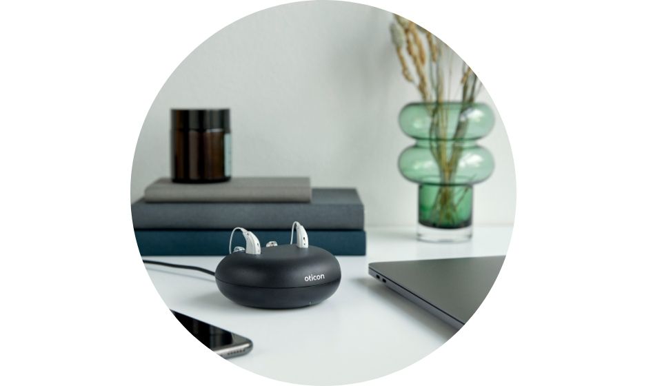 Oticon hearing aids in a charging dock placed on a white desk alongside books, a smartphone, a laptop, and decorative items.