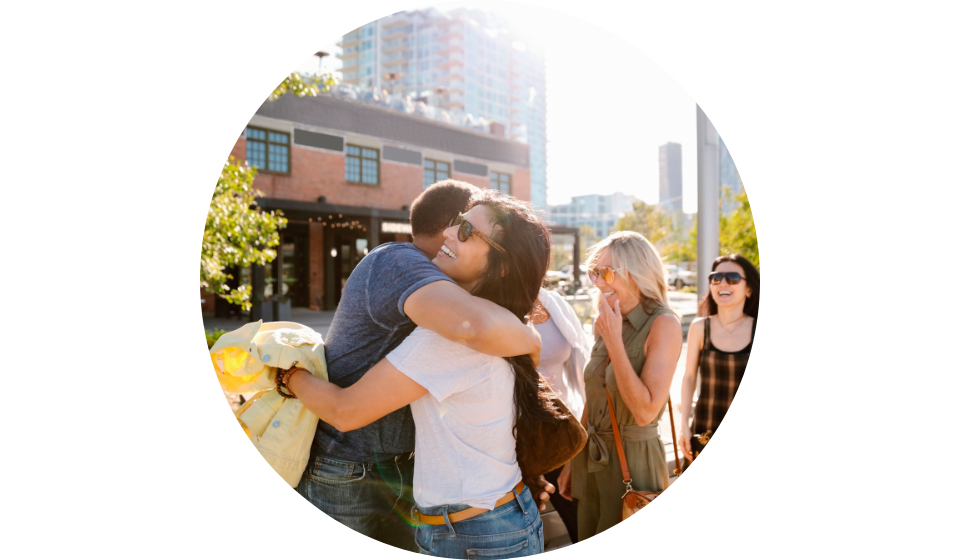 Family embracing outdoors during a sunny day, surrounded by urban buildings and greenery.