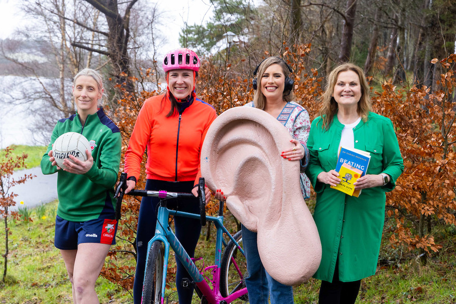 Four women stand outdoors on a lakeside path surrounded by autumnal trees. They hold objects symbolizing diverse interests: a Gaelic football, a bicycle, a large ear model, and a book titled ''Beating Brain Fog''. One wears headphones, emphasizing hearing care or awareness.