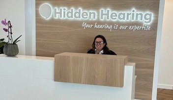 [GEN] A modern hearing clinic reception features a desk, a potted orchid, and a backdrop displaying the Hidden Hearing logo with the tagline 'Your hearing is our expertise' in illuminated text.