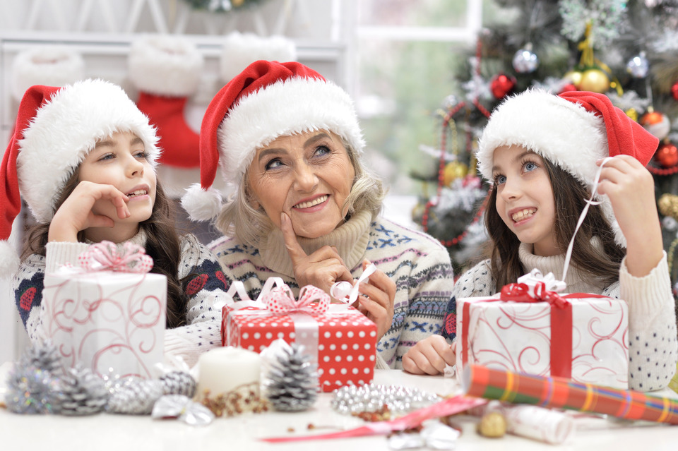Three people wearing Santa hats unwrap gifts at a festive table adorned with wrapped presents, pinecones, and ornaments, against the backdrop of a decorated Christmas tree and stockings.