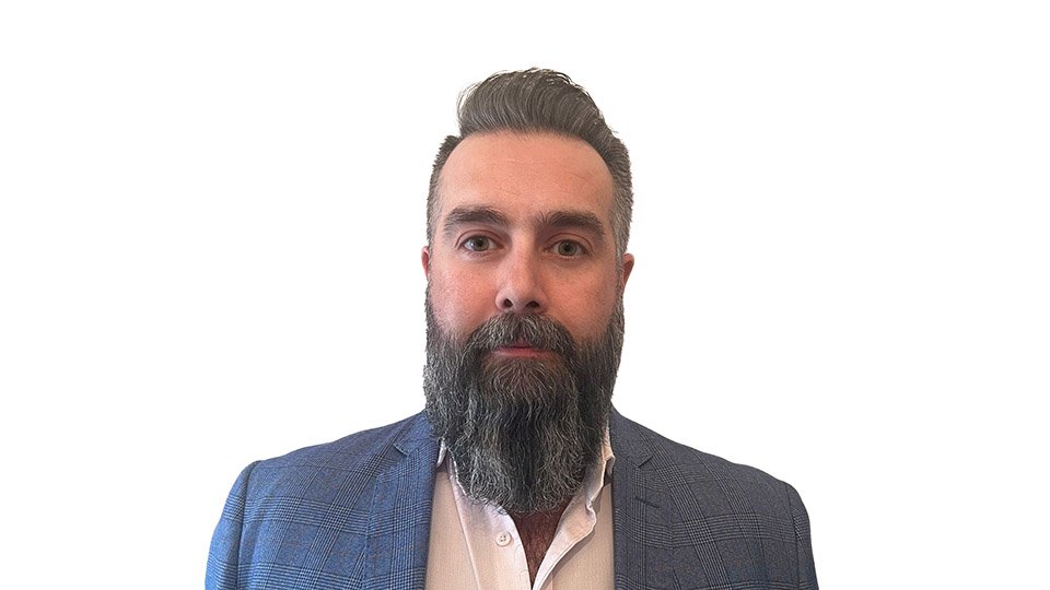 A man with a blurred face sports a full, thick beard, dressed in a blue checkered blazer and a light pink shirt, set against a plain white background. No text is visible.