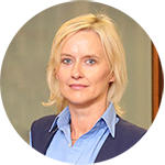 A professional woman with short blonde hair wearing a light blue shirt and navy vest stands in an indoor setting with a neutral-toned wall, likely representing a hearing clinic environment.