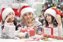 Three individuals wearing Santa hats are unwrapping festive presents in a decorated room with a Christmas tree, pinecones, and holiday-themed wrapping paper, creating a cheerful, seasonal atmosphere. No text or hearing aids are visible.