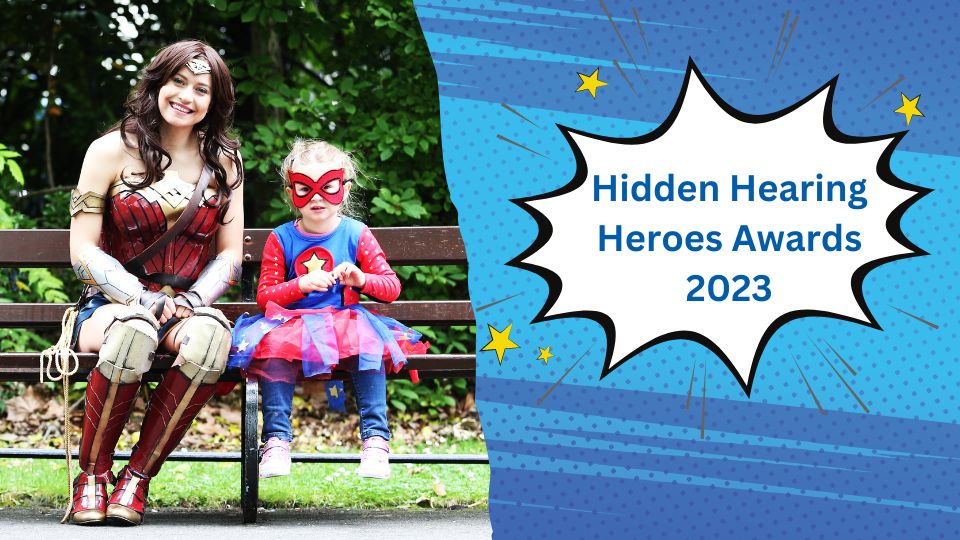 [GEN] A woman dressed as Wonder Woman and a young girl in a superhero costume sit on a park bench surrounded by greenery. Text on a comic-style blue background reads: 'Hidden Hearing Heroes Awards 2023.'