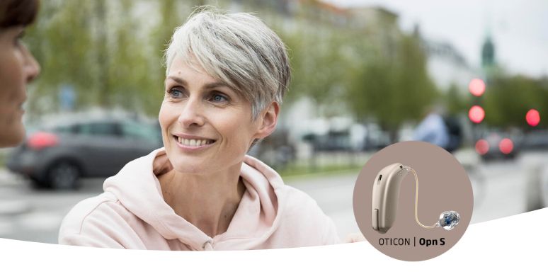A person with short gray hair speaks outdoors in an urban setting near blurred cars and trees. Inset: an Oticon Opn S hearing aid showcased with a sleek beige design.