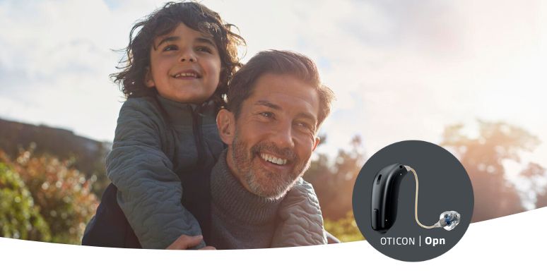 A child sits on an adult's shoulders outdoors during sunset, surrounded by blurred greenery. The image highlights an Oticon Opn hearing aid in the corner, showcasing advanced hearing care solutions.