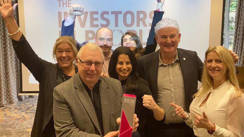 A group of people celebrates indoors, holding an award inscribed with 'INVESTORS IN PEOPLE.' Behind them is a projection screen displaying 'THE INVESTORS IN PEOPLE' text in a professional setting.