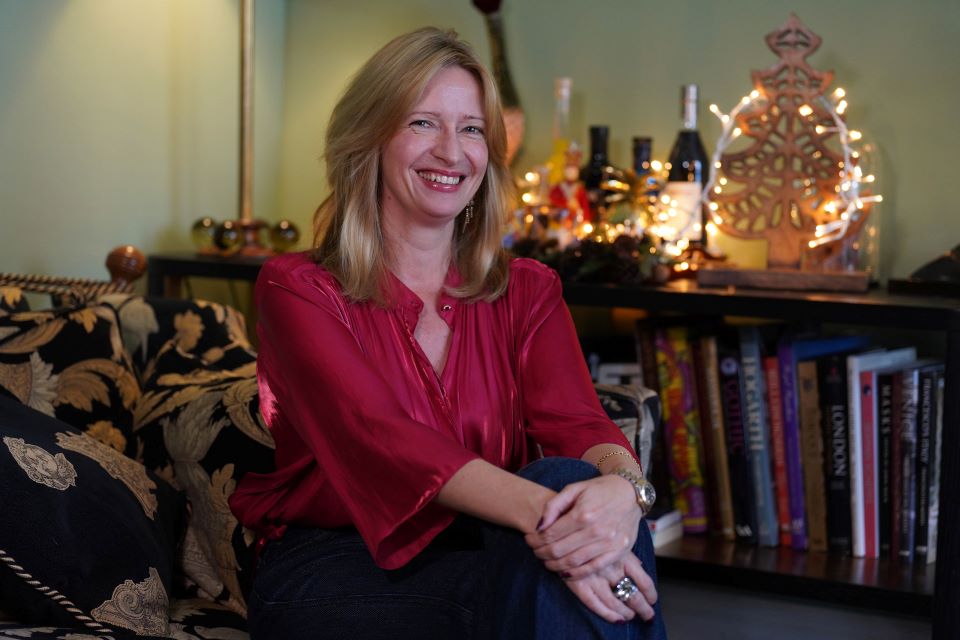 Woman in festive red blouse sitting on a patterned sofa with a decorative bookshelf and illuminated Christmas decorations in the background.