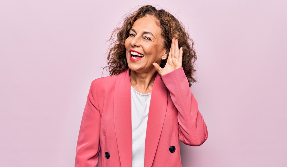 Hearing awareness concept with a woman cupping her hand to her ear while wearing a pink blazer against a pastel background.