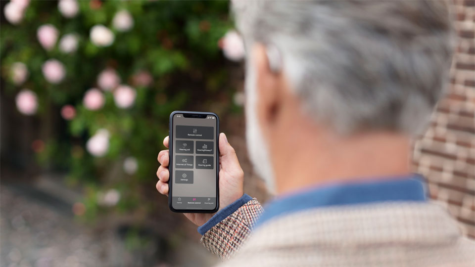 Man with gray hair using a smartphone to adjust Bluetooth hearing aids outdoors