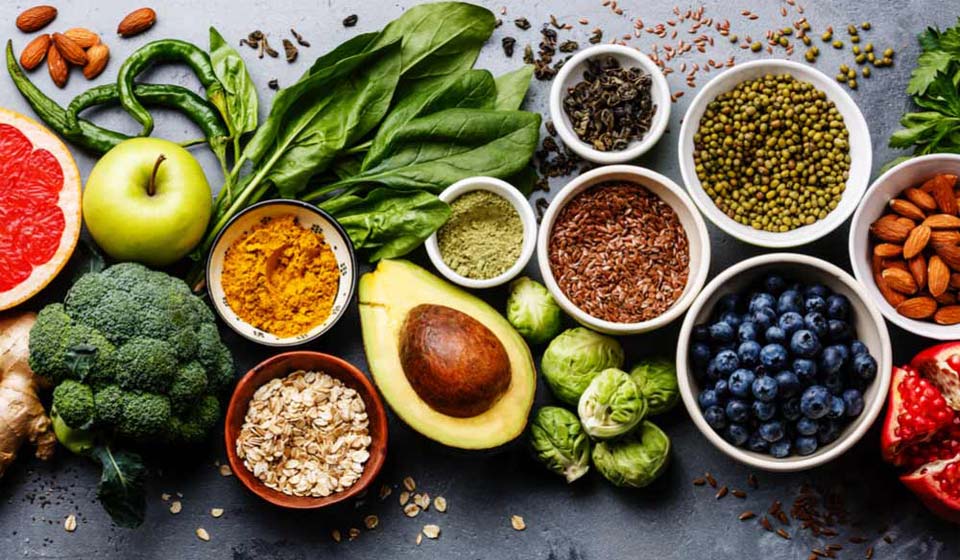 Colorful assortment of healthy foods including fruits, vegetables, nuts, seeds, and grains such as avocado, blueberries, spinach, almonds, and oats.